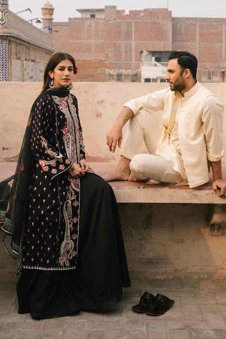 A woman in a black embroidered traditional outfit with pink floral details and a man in a cream-colored kurta and trousers, sitting on a stone ledge in an outdoor urban setting.