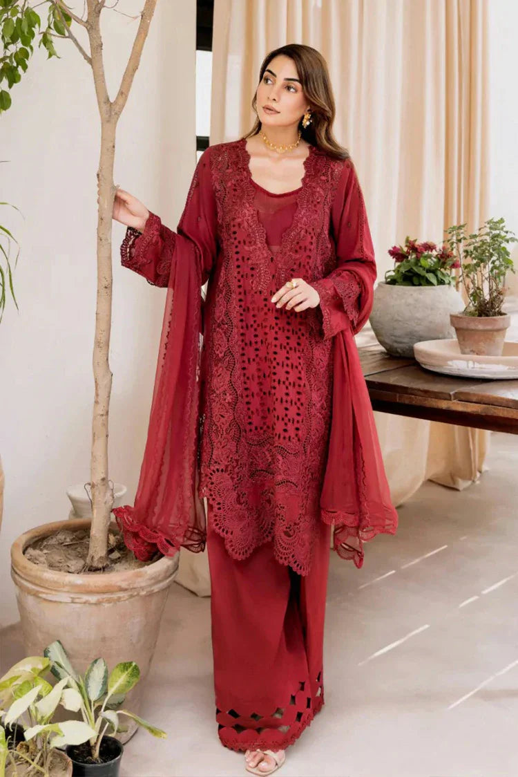 A woman models a three-piece maroon red ethnic suit set featuring a long-sleeve embroidered kurta, matching wide-leg trousers, and a sheer embroidered dupatta, set against a warm indoor background with potted plants.