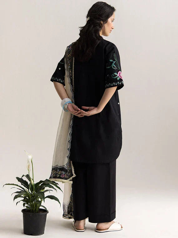 Woman wearing a black outfit with floral embroidery, standing next to a potted plant on a plain background