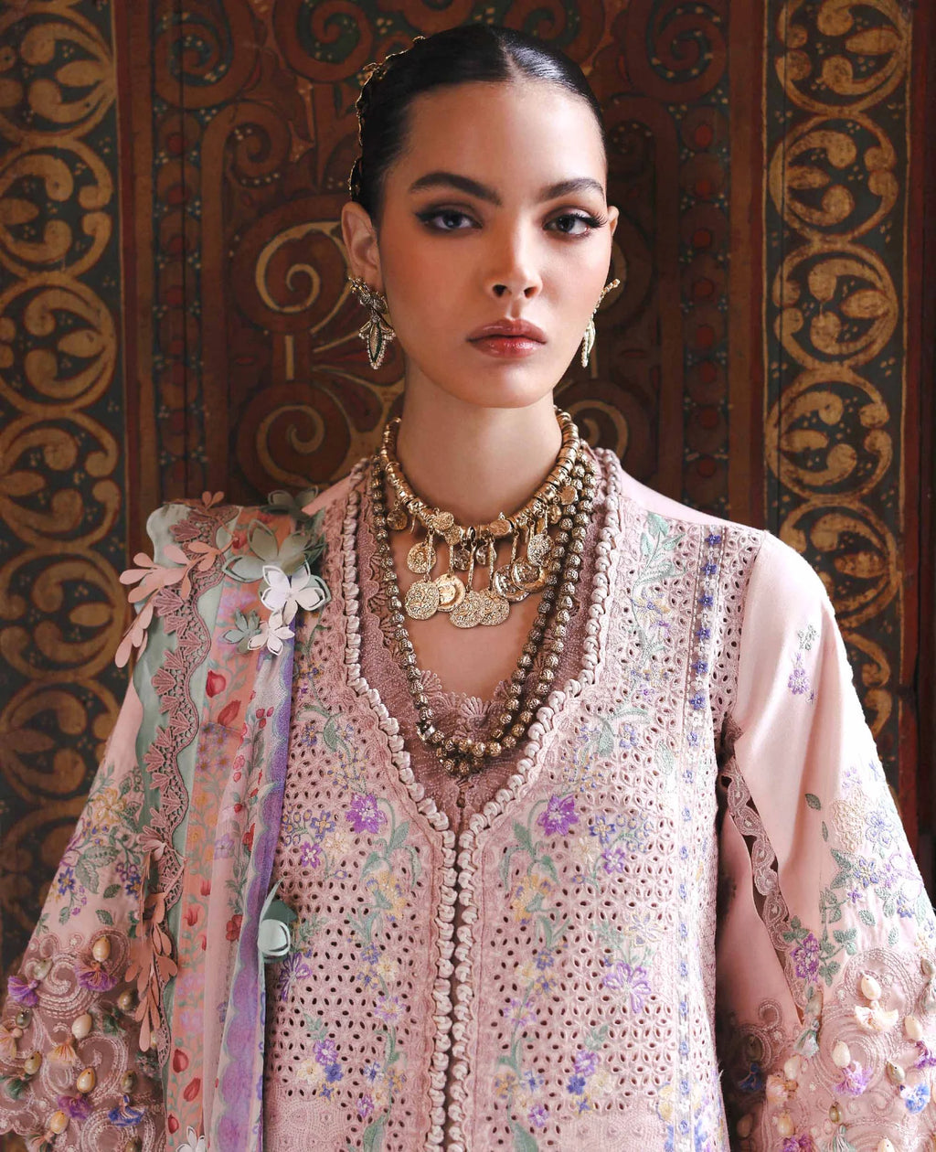 Woman wearing an ornate traditional outfit with intricate patterns and jewelry against a decorative background.
