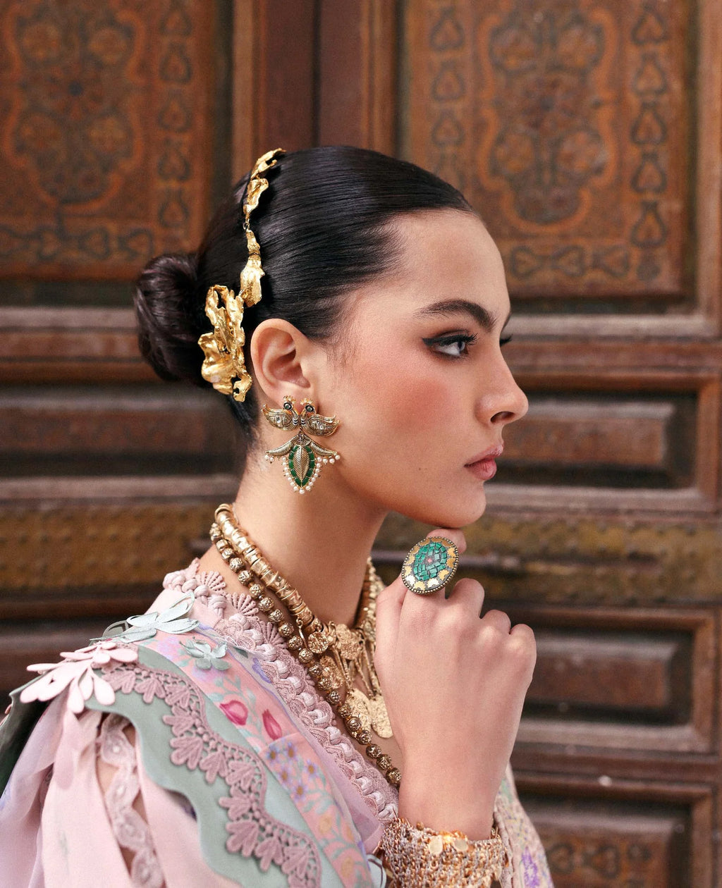 Woman wearing traditional jewelry and a pink outfit in front of an ornate wooden background