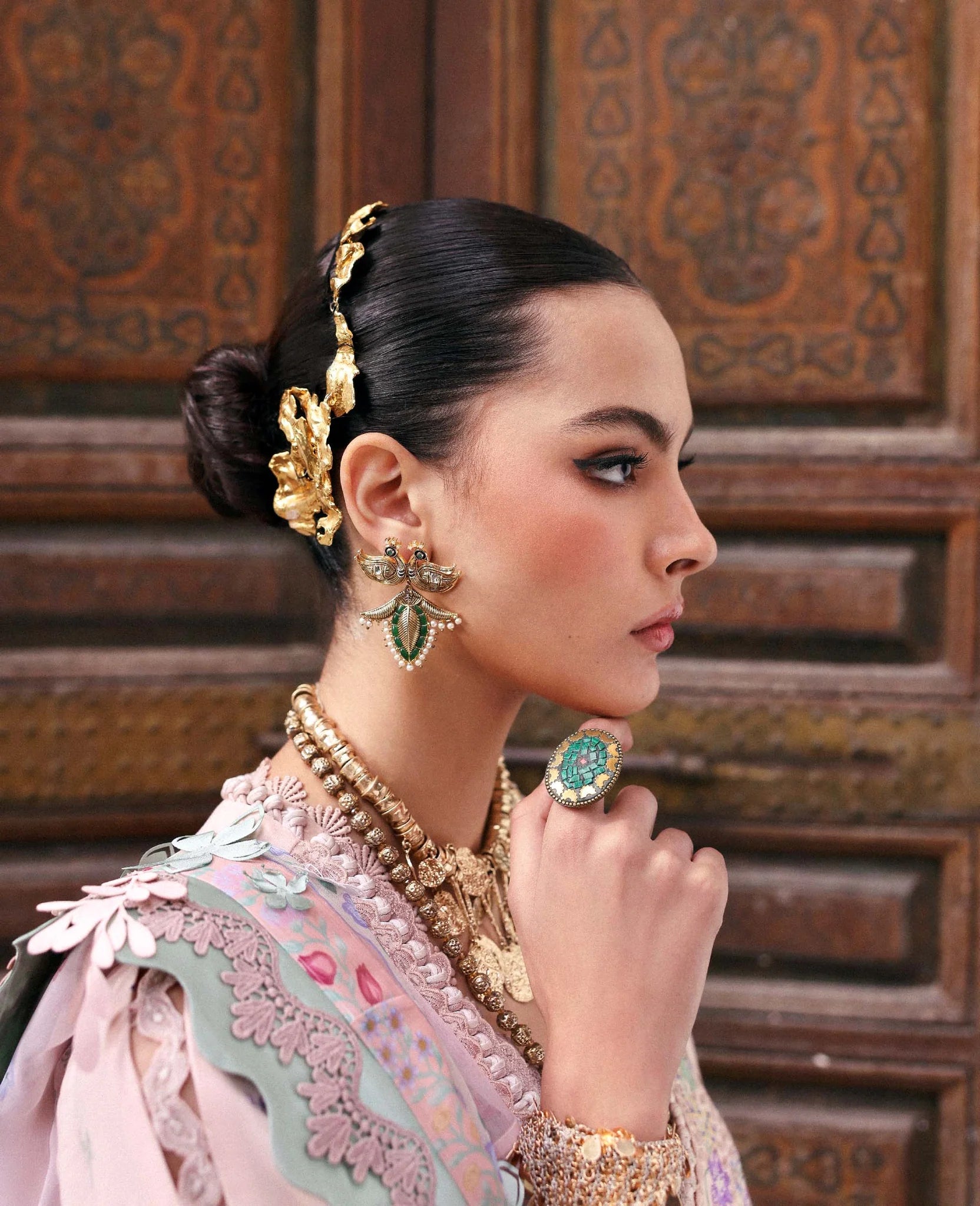 Woman wearing traditional jewelry and a pink outfit in front of an ornate wooden background