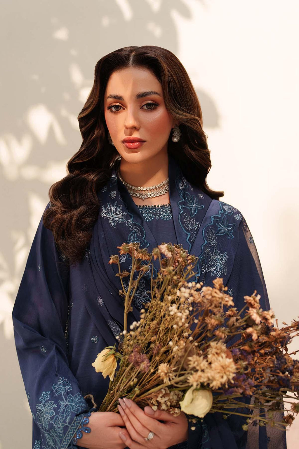 Woman in a dark blue embroidered outfit holding dried flowers against a neutral background