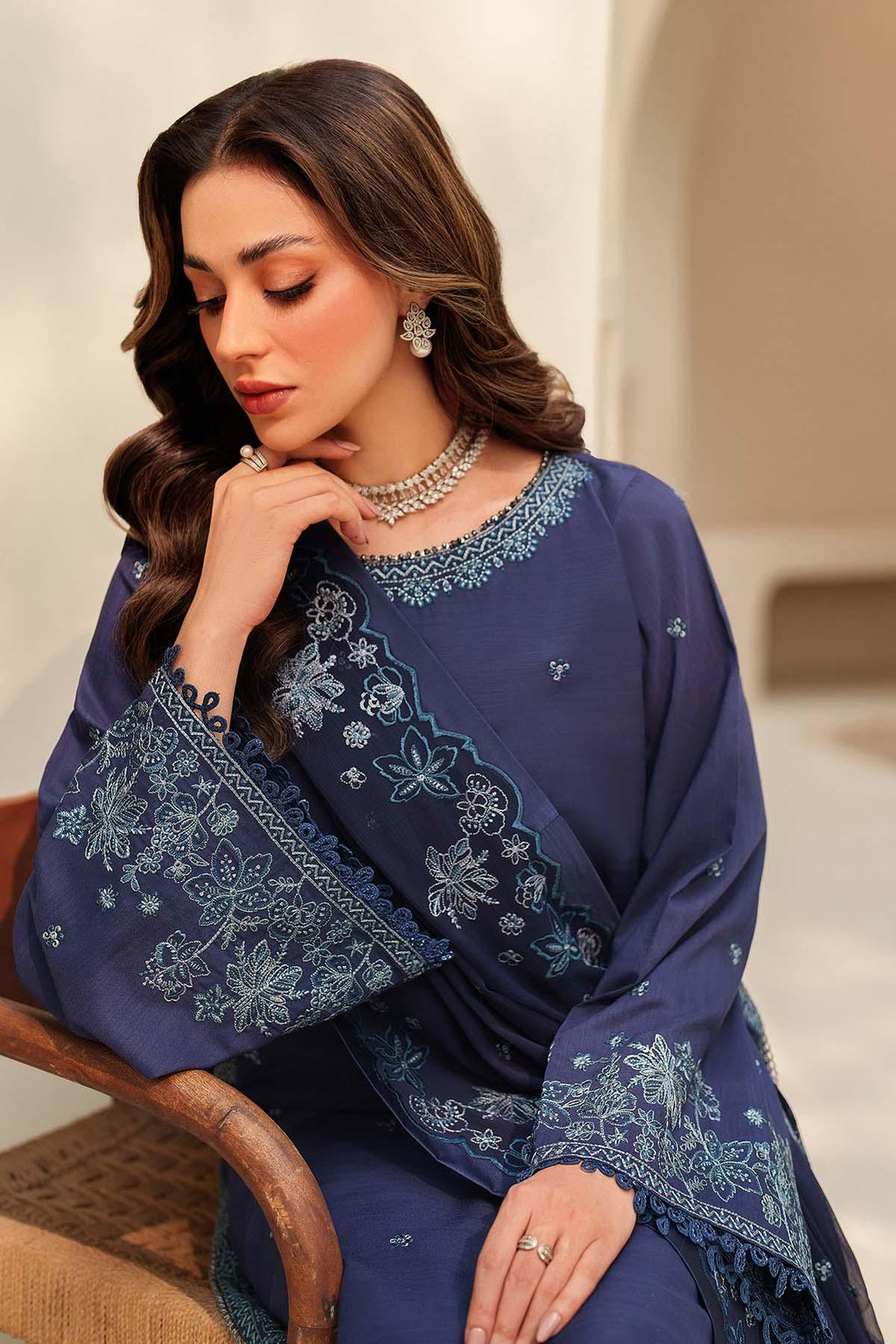 Woman wearing a blue embroidered outfit sitting on a wooden chair.