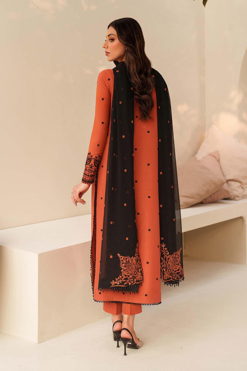 Woman wearing an orange and black traditional outfit with a matching dupatta in a minimalistic setting.