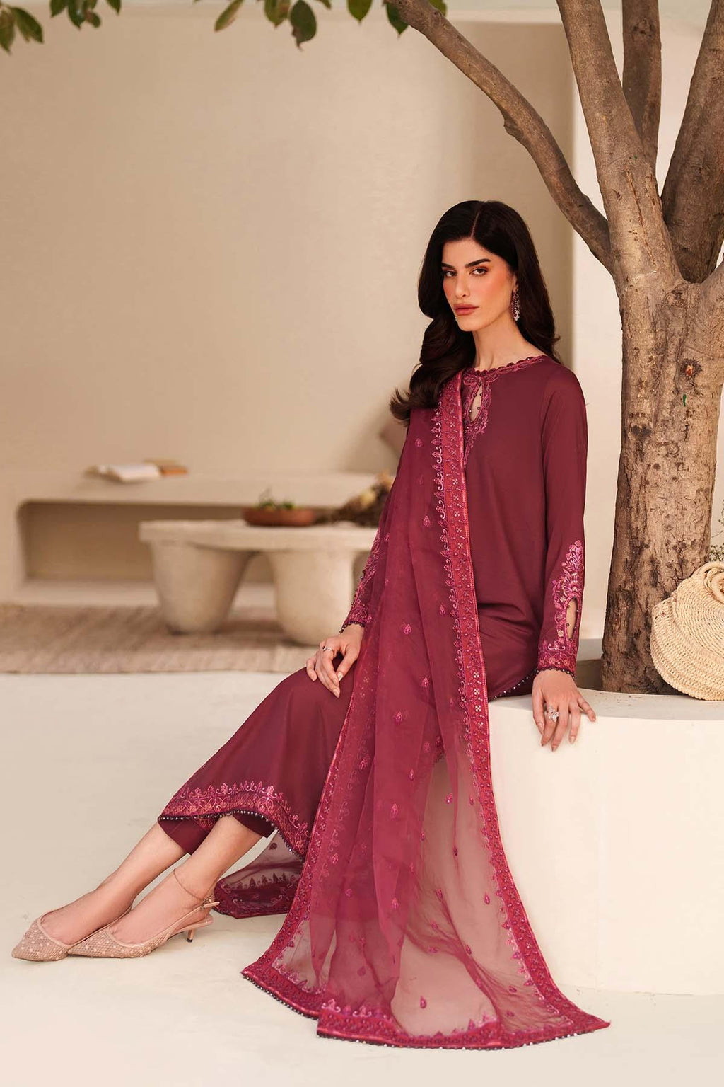 Woman in a maroon traditional outfit with intricate designs, standing in a minimalistic indoor setting.