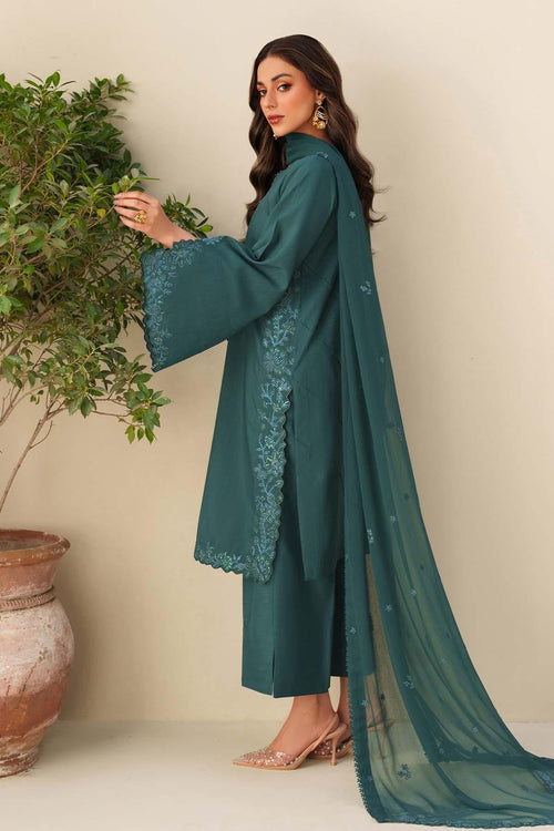 Woman in a teal traditional outfit with floral embroidery, standing next to a potted plant.