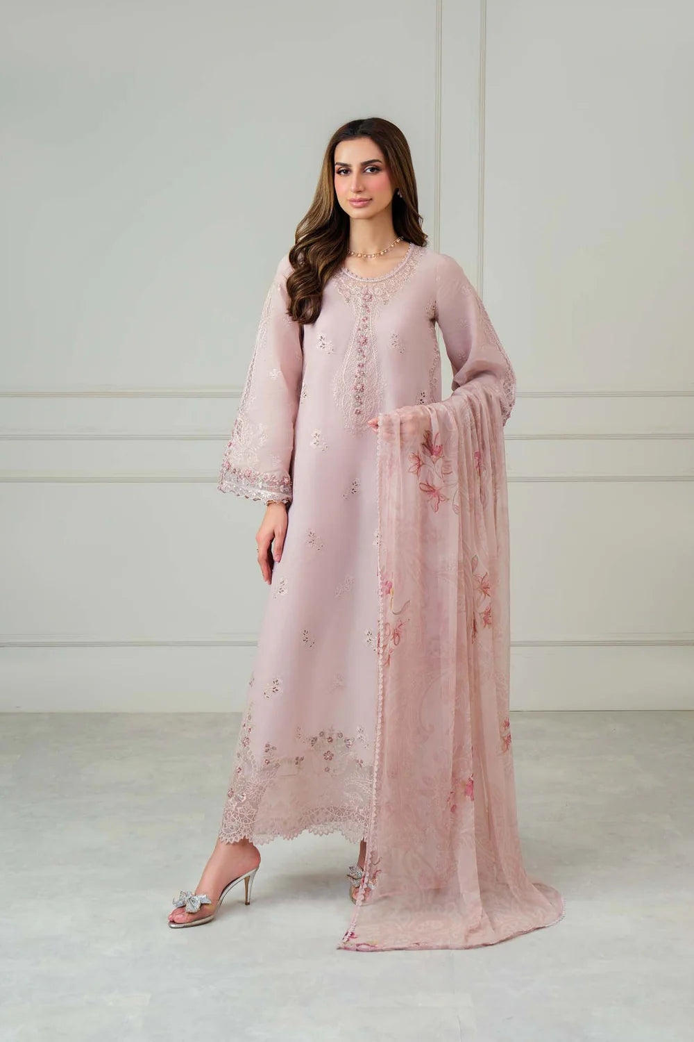 A woman models a light pink traditional outfit consisting of a long kurta with floral embroidery, matching flared sleeves, and a sheer dupatta draped over one arm, set against a neutral studio background.