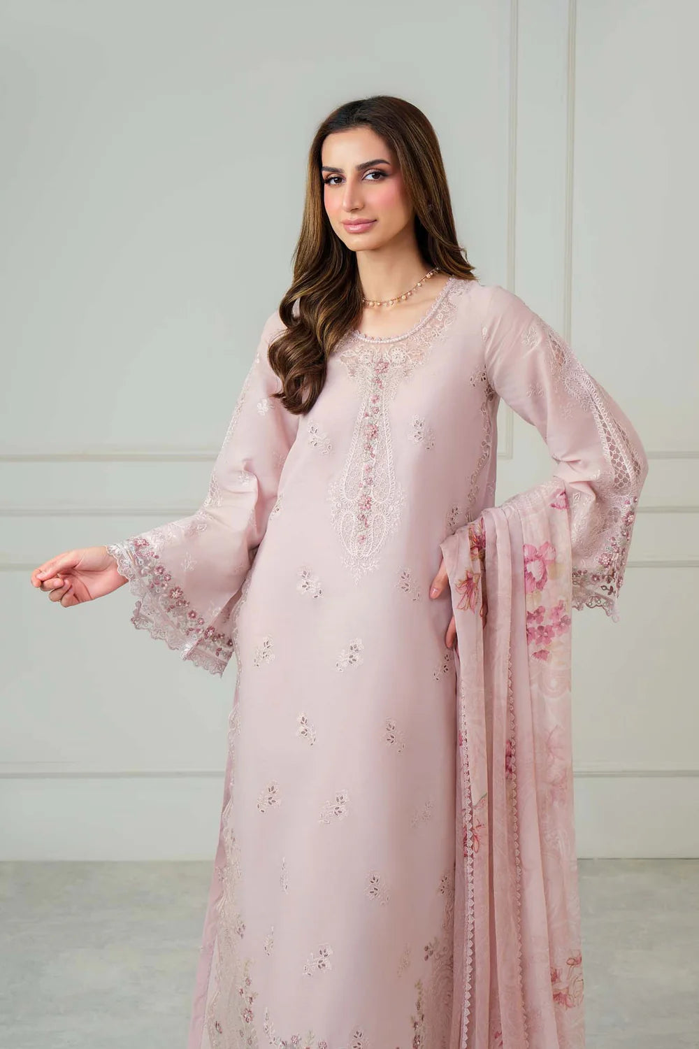 A woman modeling a light pink traditional outfit with matching dupatta, featuring embroidered details and sheer bell sleeves, set against a neutral indoor background.