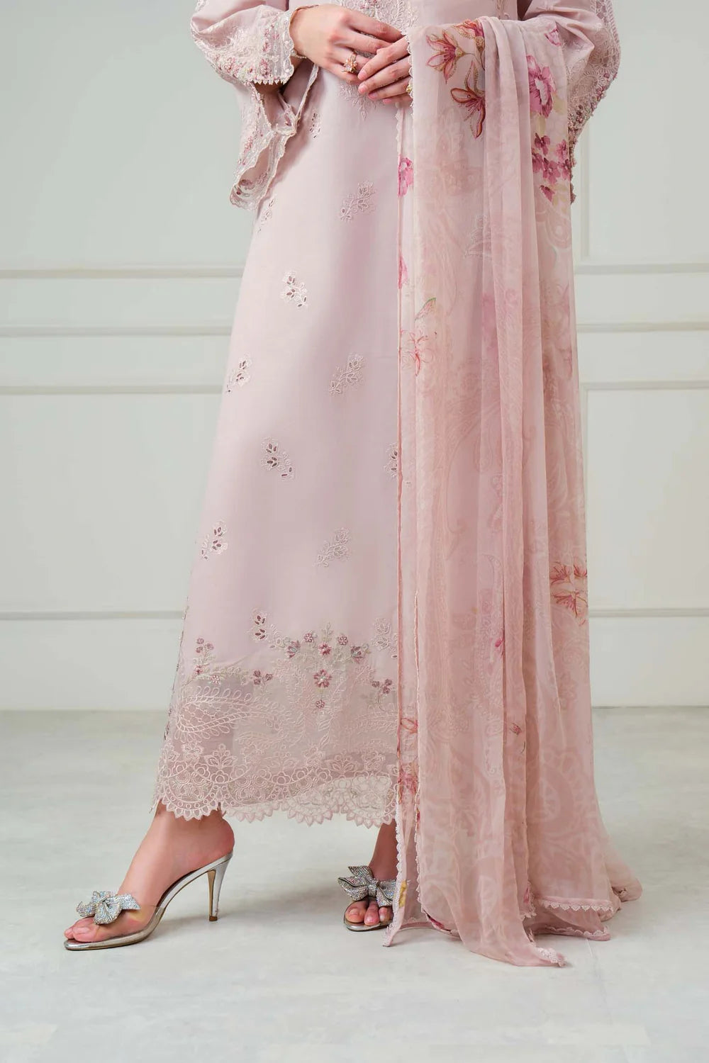 A model wearing a light pink traditional outfit with floral embroidery and a matching sheer dupatta, paired with clear high-heeled sandals.