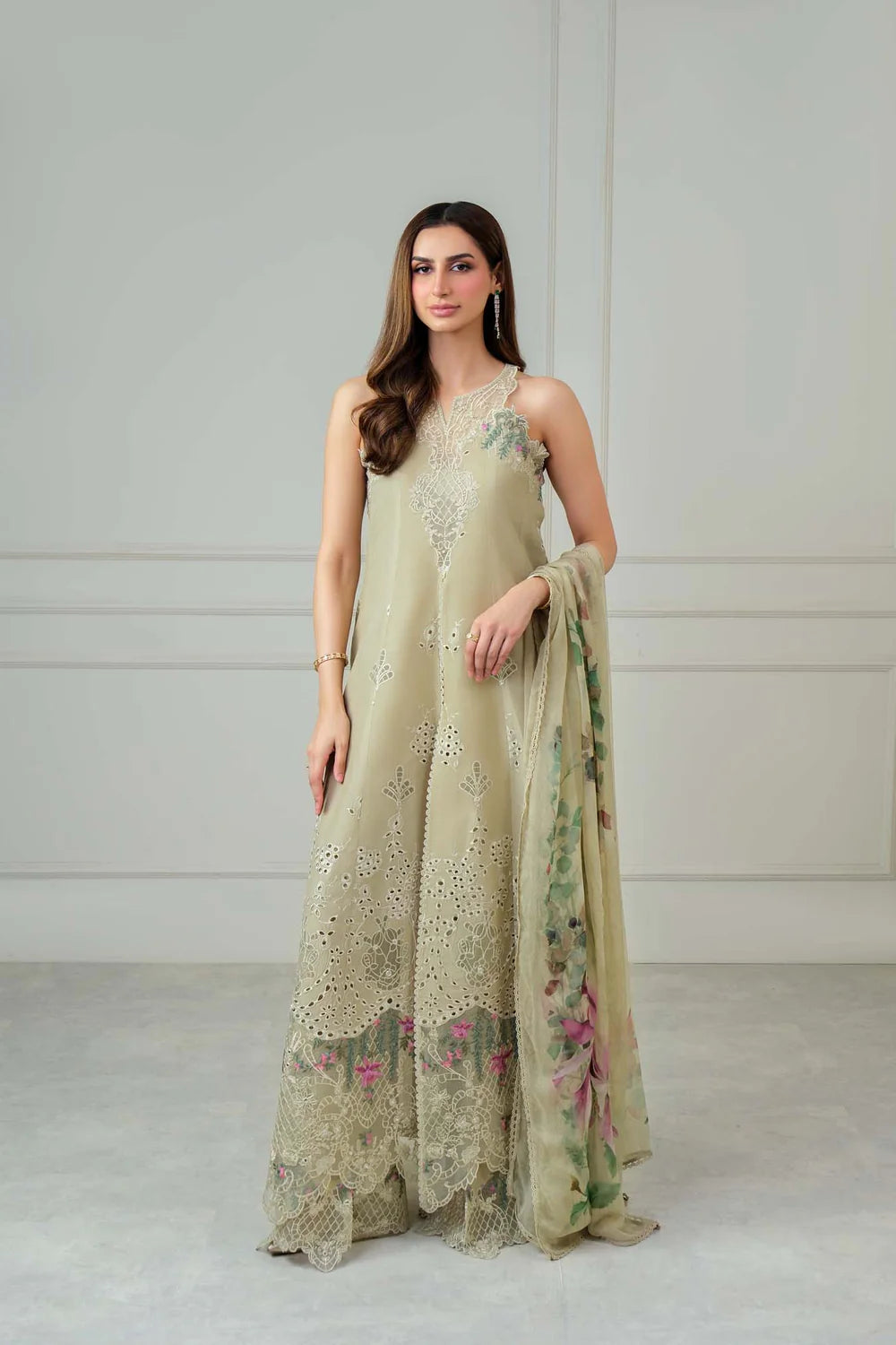 A woman models a sleeveless beige anarkali gown with intricate floral embroidery and a matching sheer dupatta featuring a green and pink floral print, set against a neutral studio background.