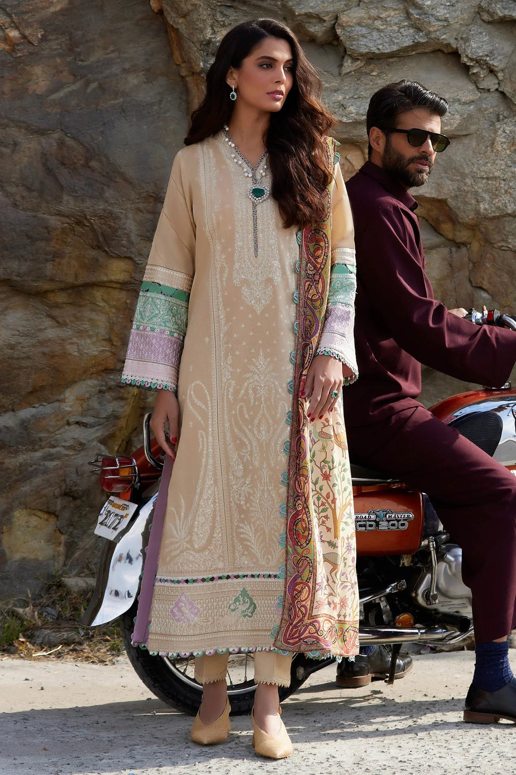 Woman in traditional outfit with man on a motorcycle against a stone wall.