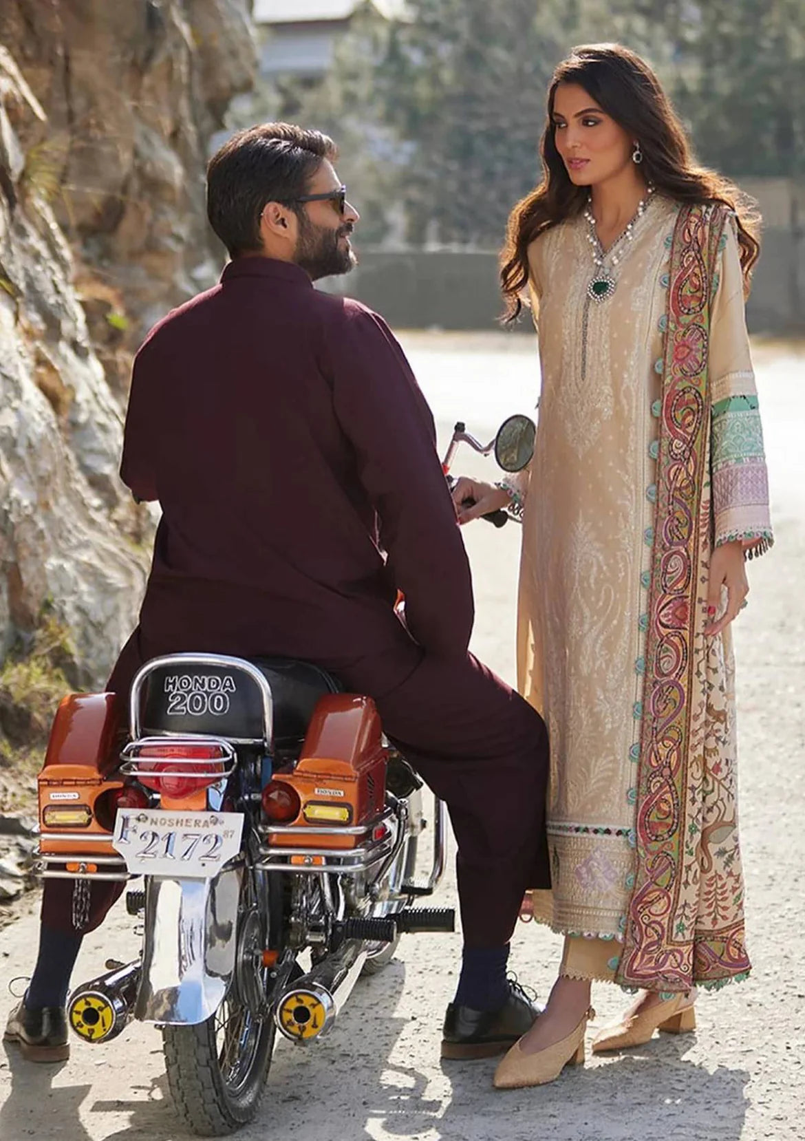 A man in a dark plum kurta sits on a Honda Zorro motorcycle next to a woman wearing a beige embroidered traditional outfit with a multicolor paisley border, set against a rocky outdoor background.