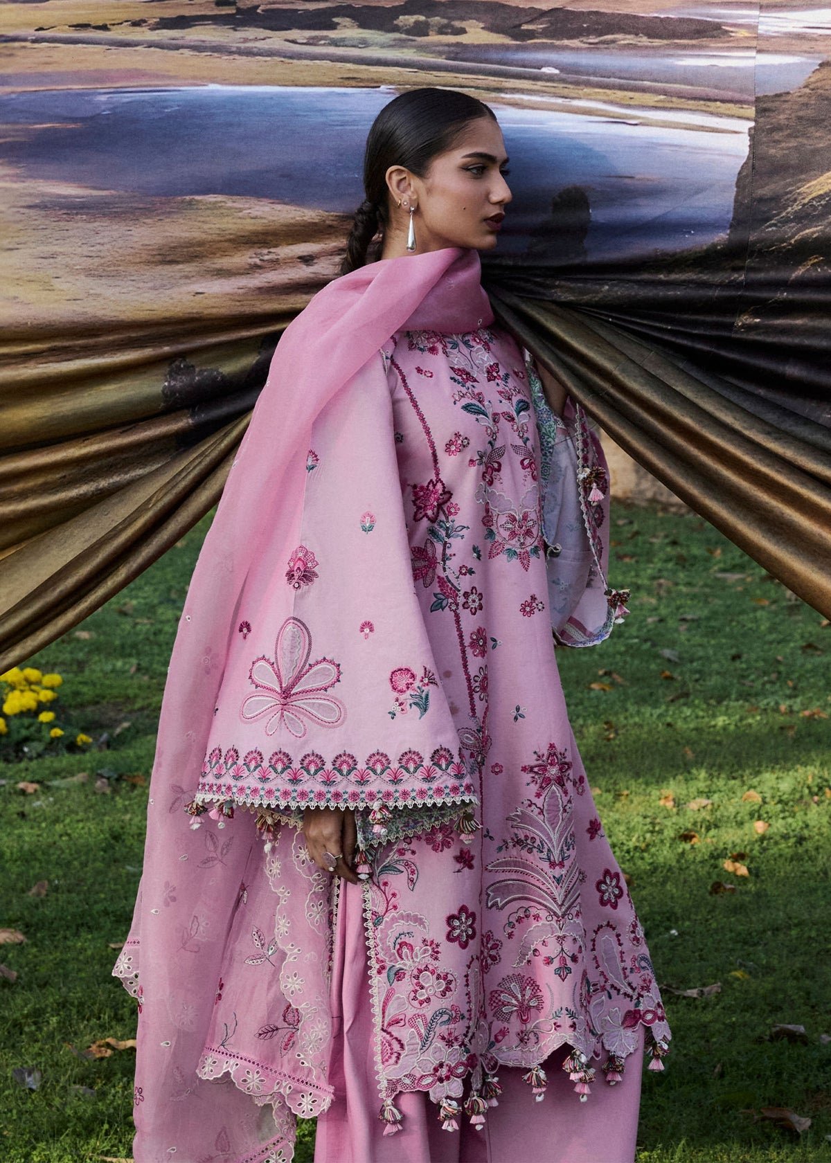 Woman in a pink embroidered outfit standing in a natural setting with large cylindrical objects in the background.
