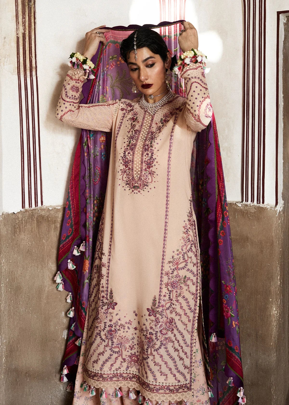 Woman wearing a beige embroidered dress with a colorful dupatta against a textured wall.