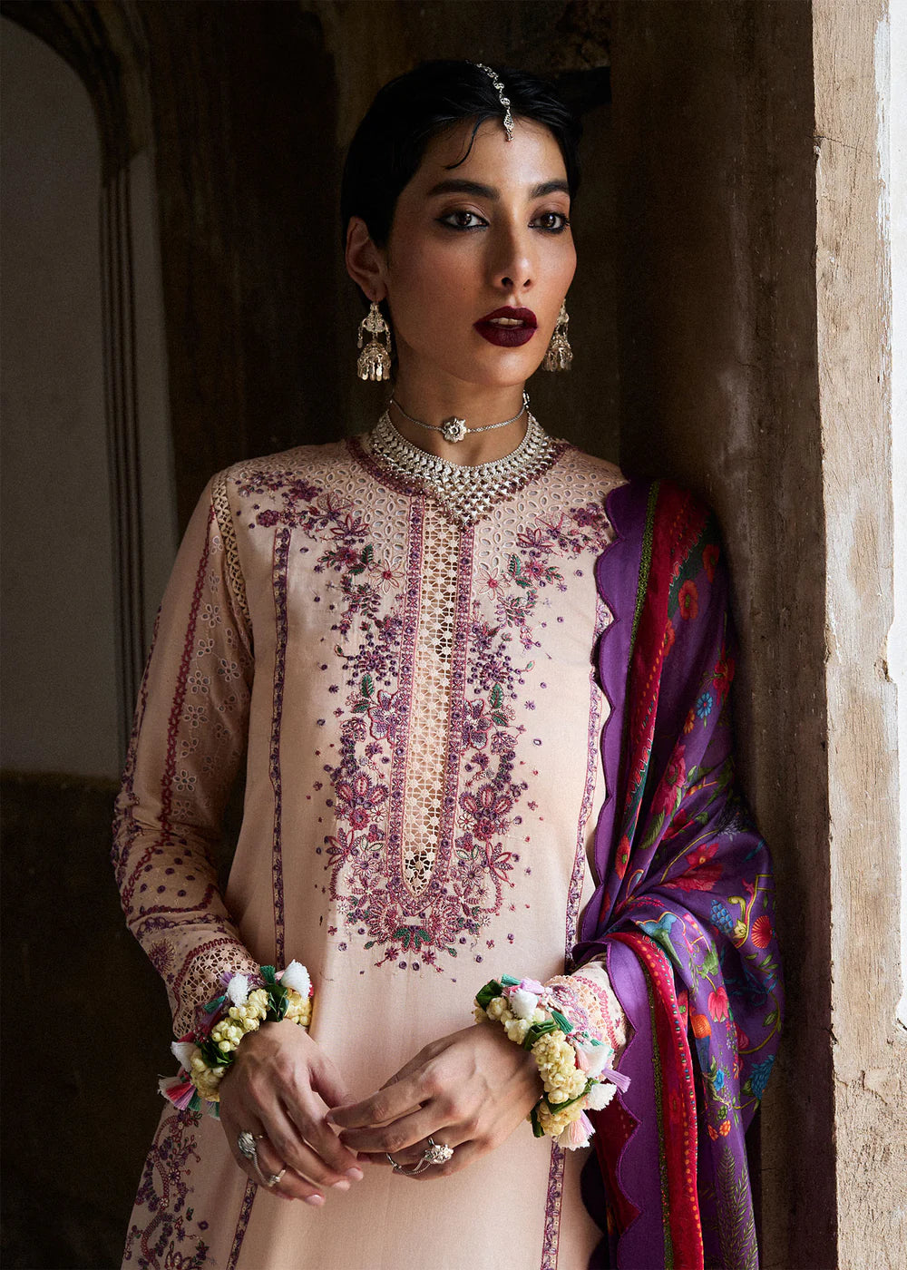 Woman in traditional attire with intricate embroidery and jewelry, standing against a textured wall.