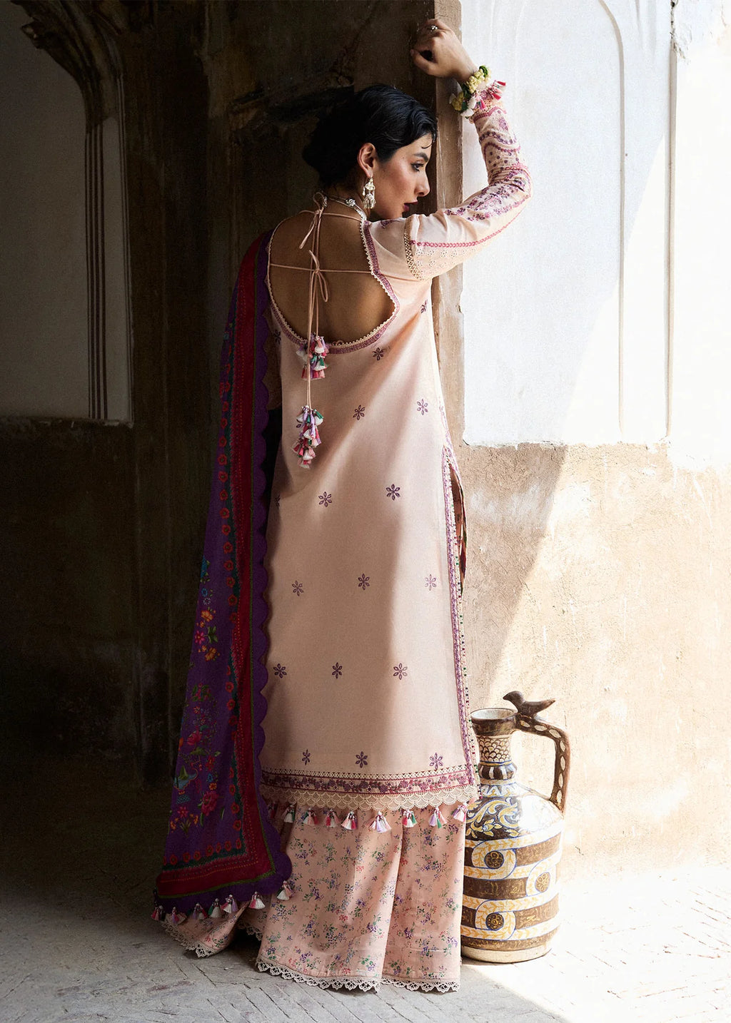Woman in a traditional outfit standing near a window with a decorative vase.