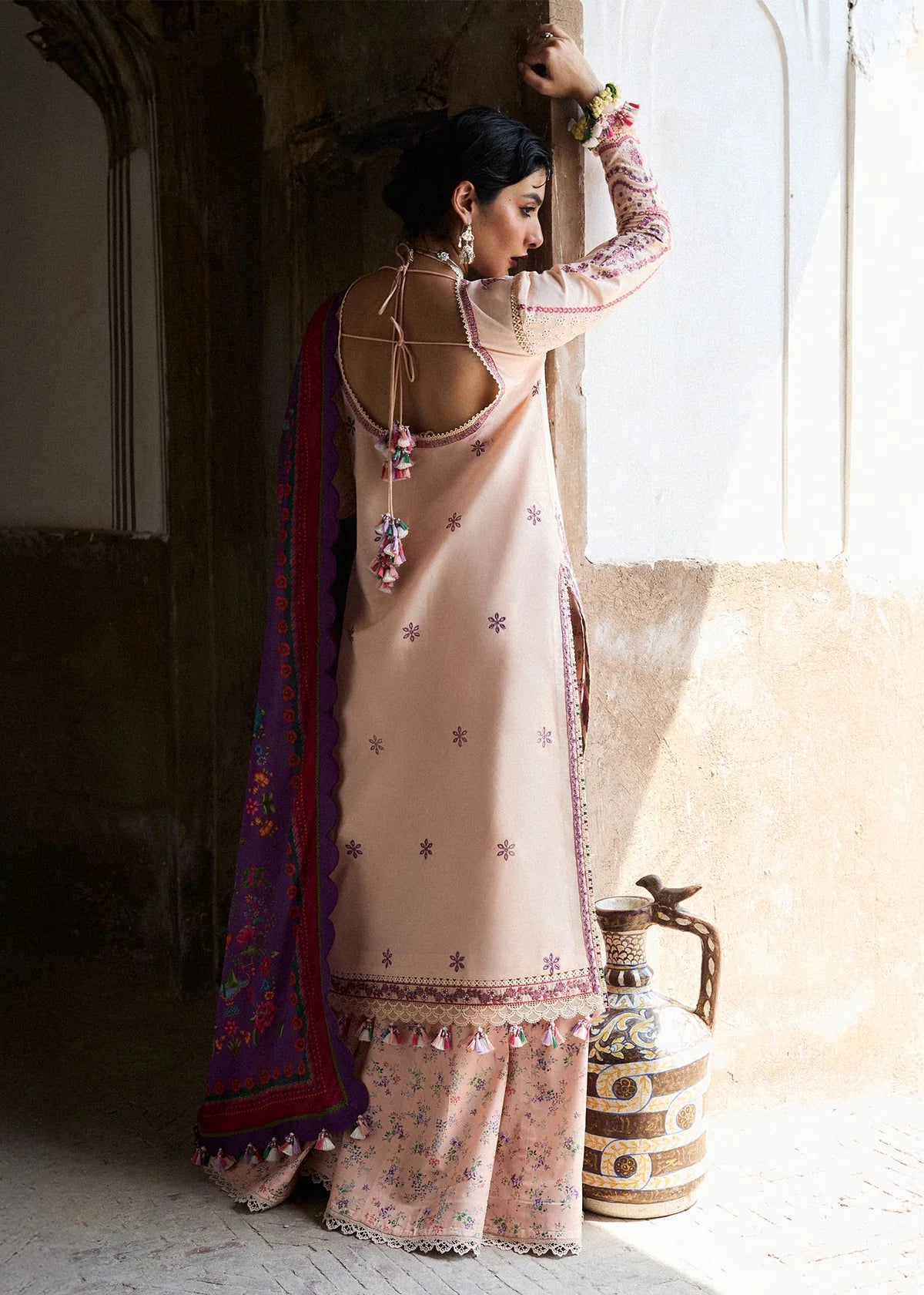 Woman in a traditional outfit standing near a window with a decorative vase.