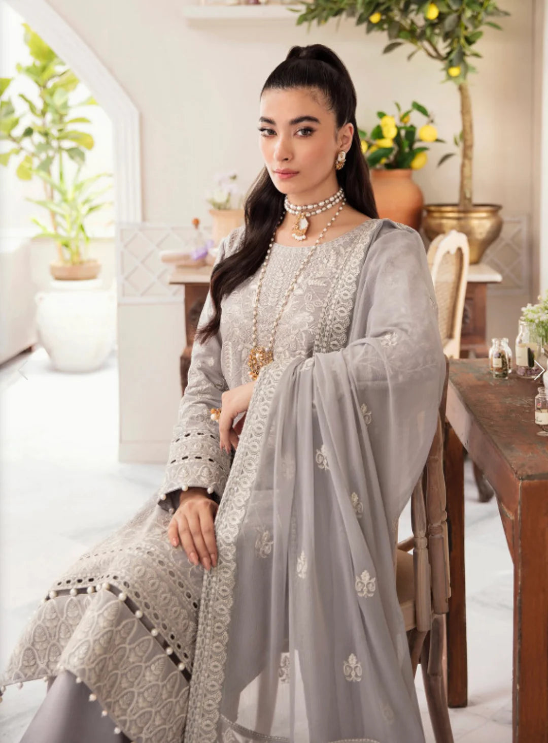 A woman seated indoors wearing a light gray traditional outfit with intricate embroidery and a matching sheer dupatta draped over her lap. The setting includes a wooden table, chairs, and potted plants in a bright interior.