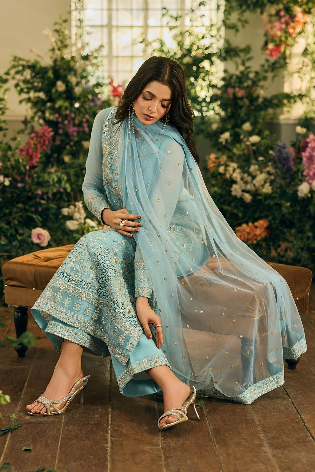 Woman in a light blue traditional outfit with a sheer dupatta, sitting on a bench with flowers in the background.