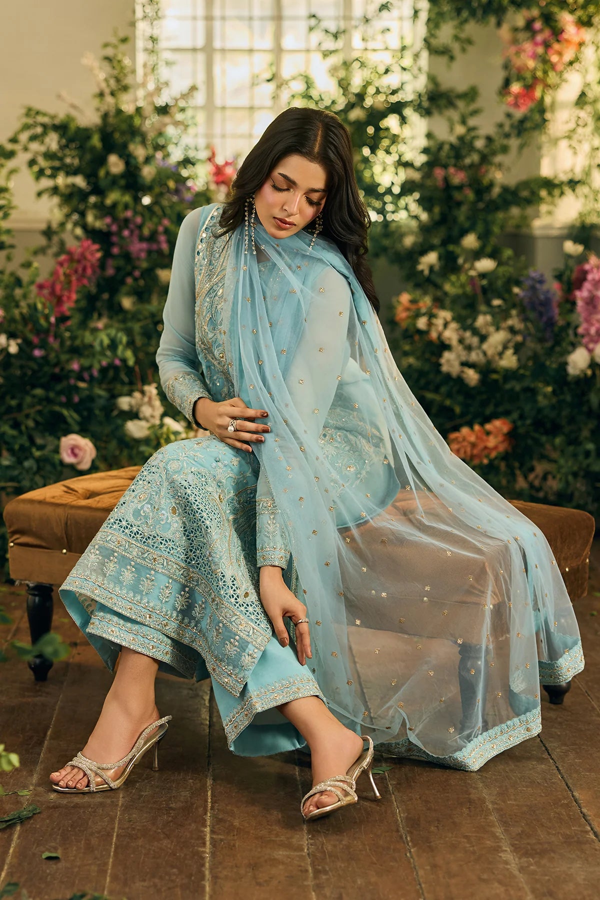 Woman in a light blue traditional outfit with a sheer dupatta, sitting on a bench with flowers in the background.