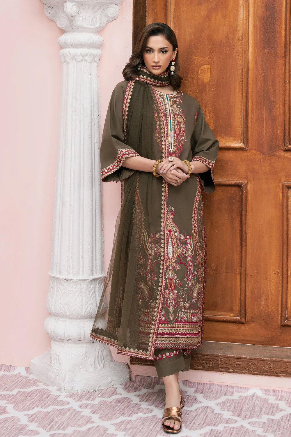 A woman models an olive green traditional kurta set with intricate red and gold embroidery, standing in front of a pink wall and wooden door.