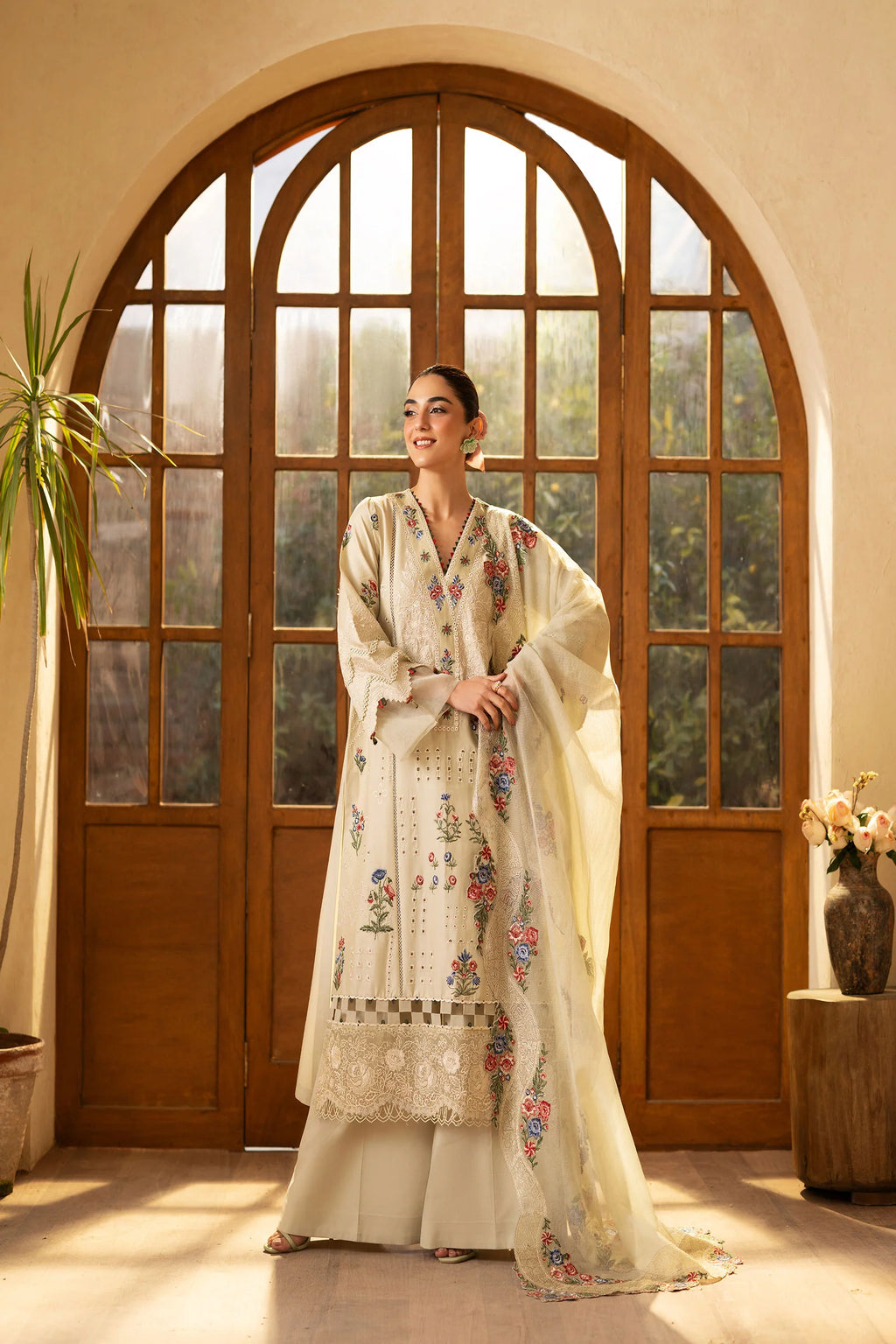 Woman in a traditional outfit standing in a room with large wooden doors and a plant.