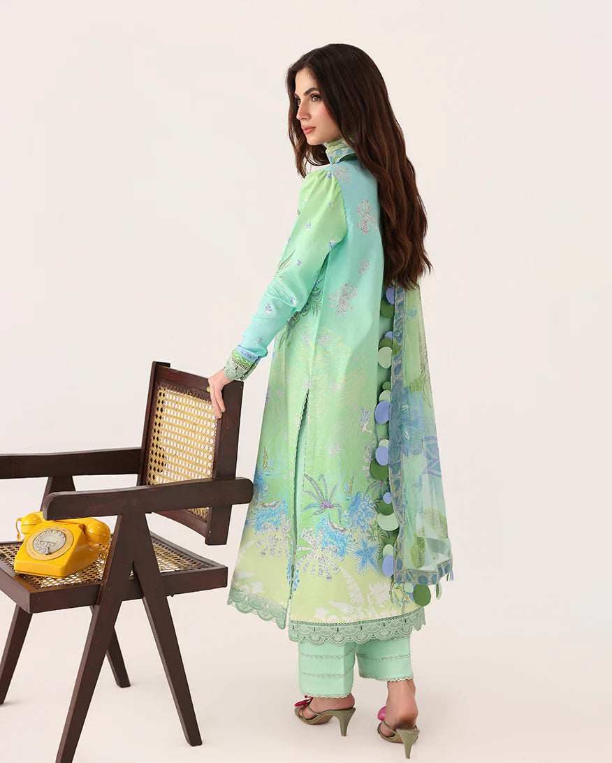 A woman modeling a mint green three-piece salwar kameez suit with floral embroidery, standing next to a dark wooden chair with a yellow rotary phone on it, against a plain off-white background.