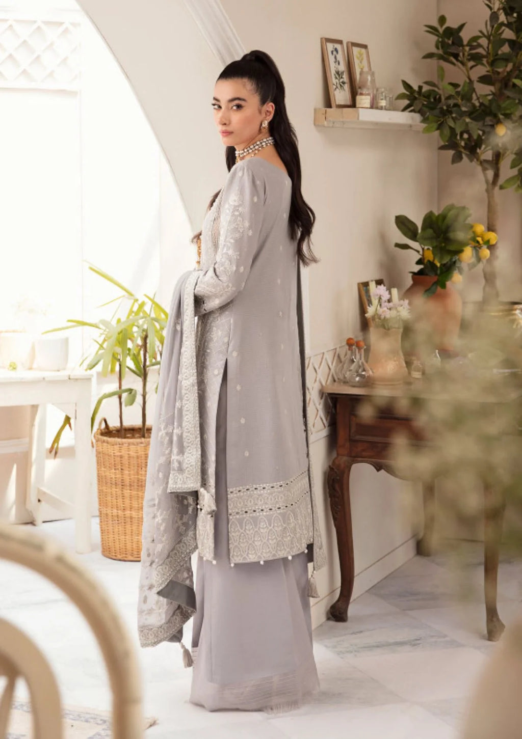 A woman models a traditional light gray three-piece outfit with matching dupatta in a bright, modern indoor setting with plants and wooden furniture.