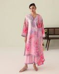 A woman modeling a pink traditional outfit with a floral print, featuring a long kurta, matching pants, and silver sandals, set against a light beige studio background with a wooden bench.