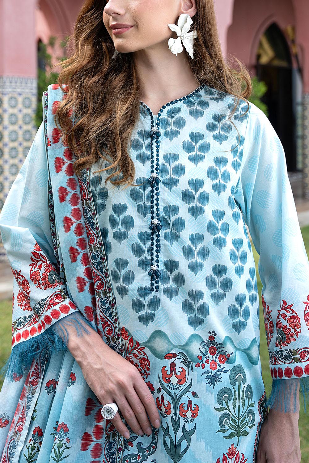 Woman wearing a light blue embroidered dress with red and green patterns, standing outdoors.