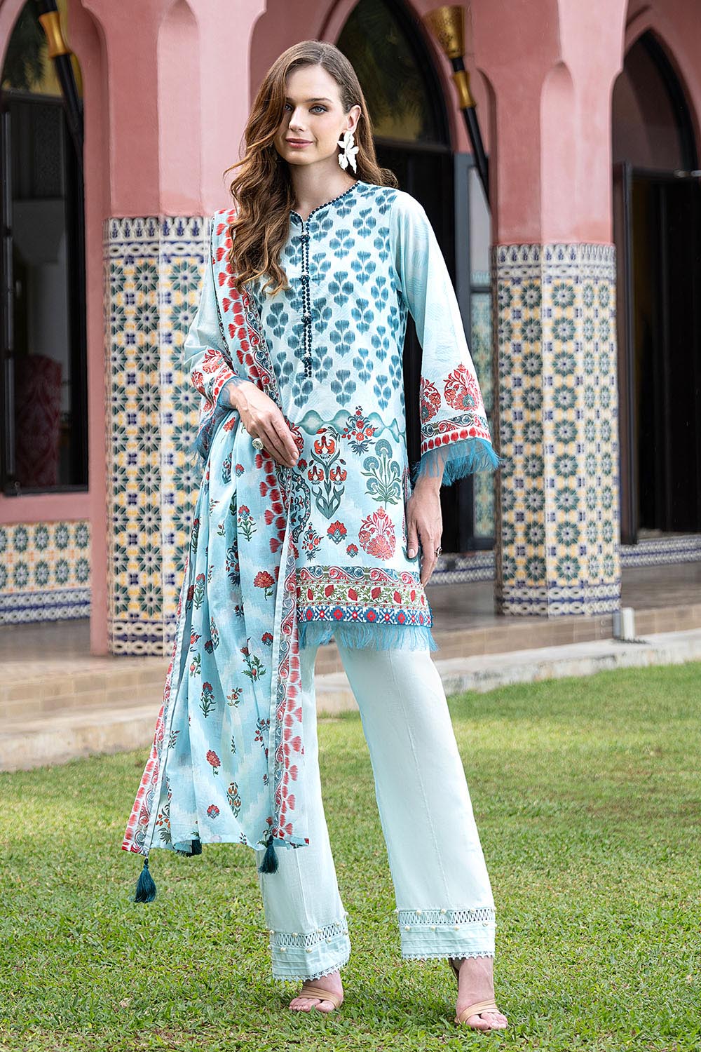 Woman in a light blue embroidered outfit standing on grass with a colorful tiled wall in the background