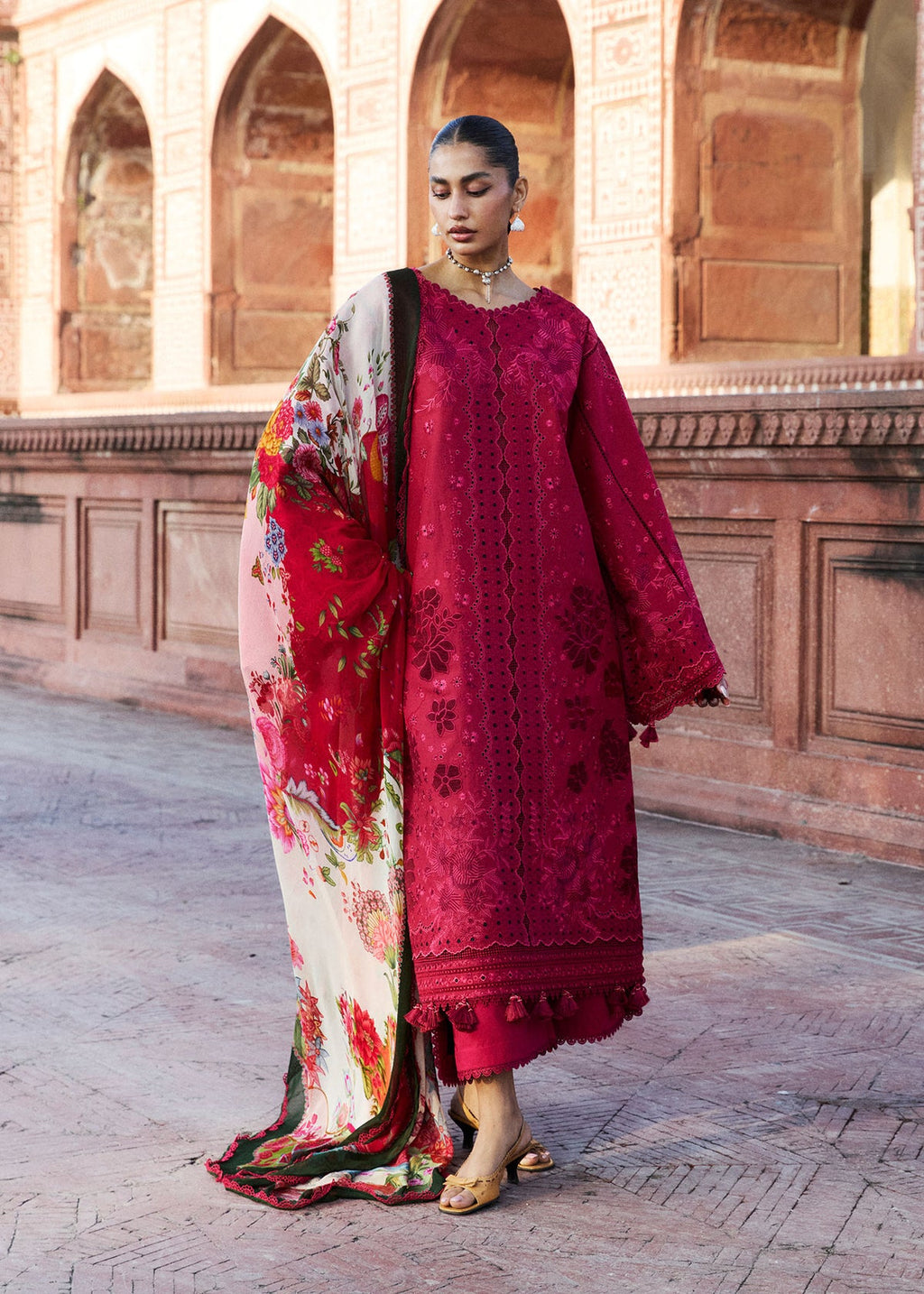 Woman in a red embroidered dress with a floral dupatta standing in front of an architectural background.