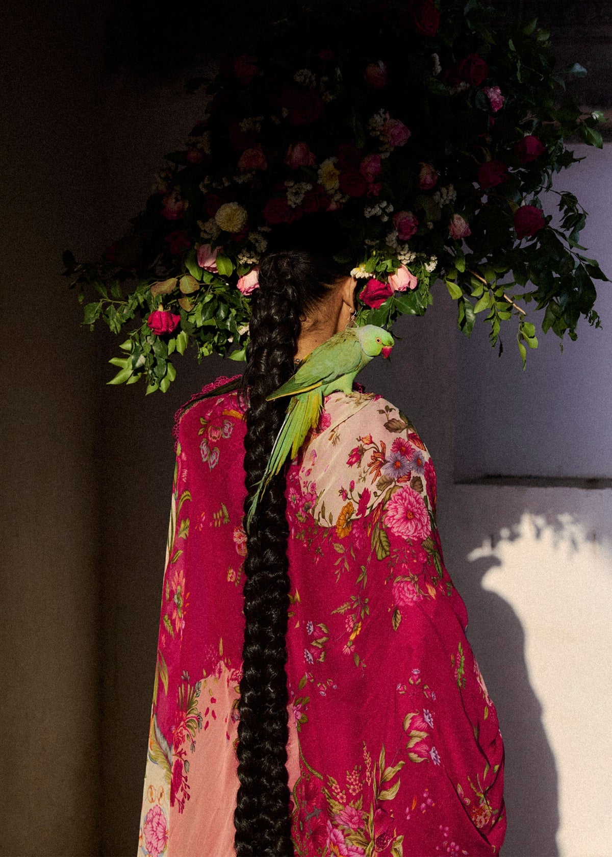 Person wearing a floral dress with a parrot perched on it, standing under a floral arch.