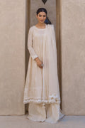 A woman modeling a traditional cream-colored three-piece outfit with a long kurta, wide-leg pants, and a matching dupatta, standing in front of a textured beige wall.