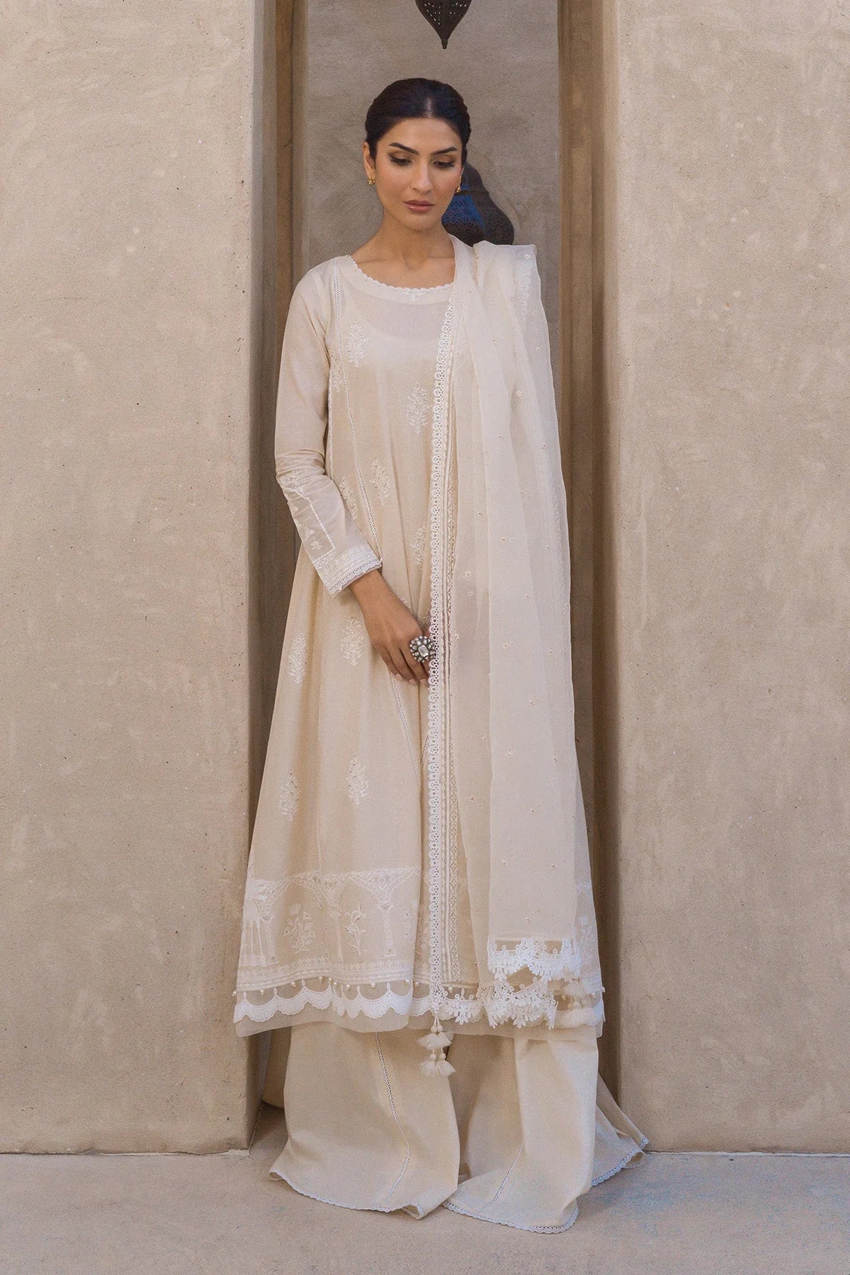 A woman modeling a traditional cream-colored three-piece outfit with a long kurta, wide-leg pants, and a matching dupatta, standing in front of a textured beige wall.