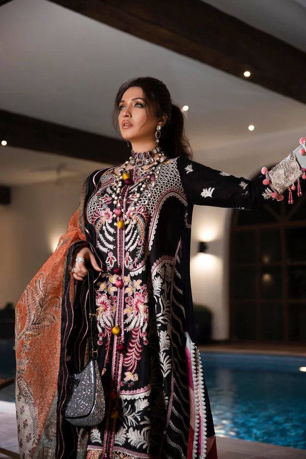 A woman models a traditional embroidered black three-piece outfit with pink, white, and gold details, featuring a matching patterned dupatta, in an elegant indoor setting with a swimming pool in the background.