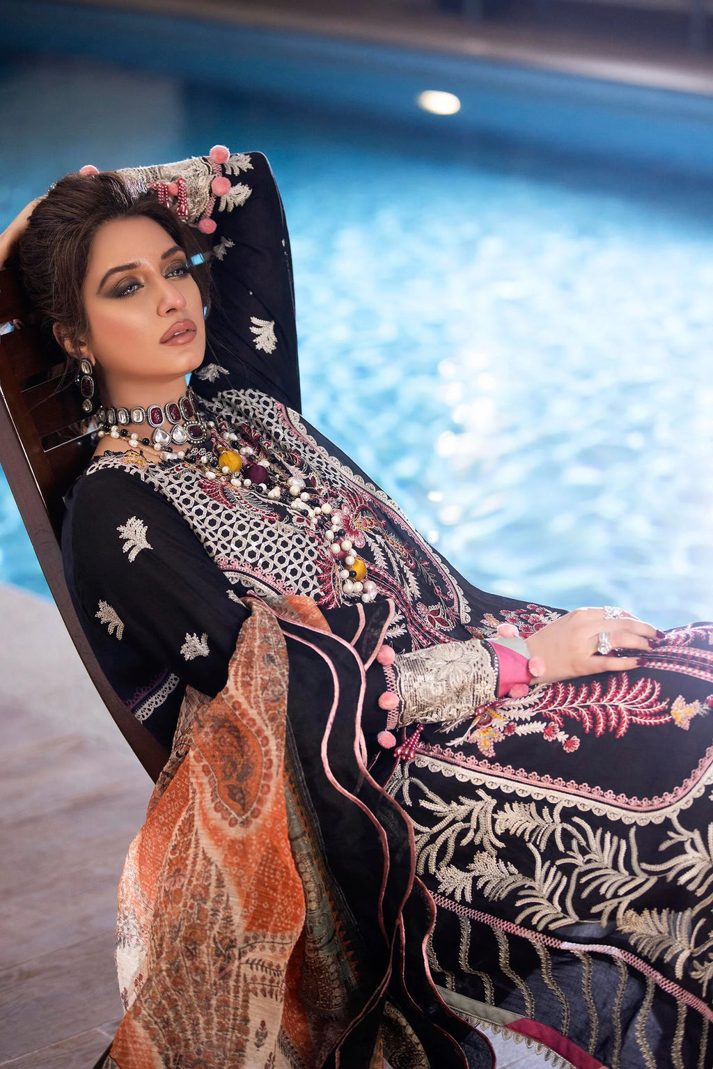A woman reclines by a swimming pool wearing an ornate black embroidered traditional outfit with intricate multicolor floral and geometric patterns, a matching dupatta, and heavy metallic jewelry.