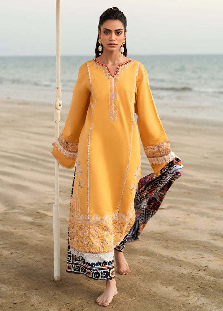 A woman stands barefoot on a sandy beach wearing a long yellow embroidered kurta with a multicolor patterned dupatta. The background shows the ocean and a pale sky.