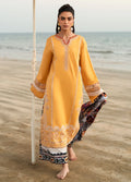 A woman stands barefoot on a sandy beach wearing a long yellow embroidered kurta with a multicolor patterned dupatta. The background shows the ocean and a pale sky.