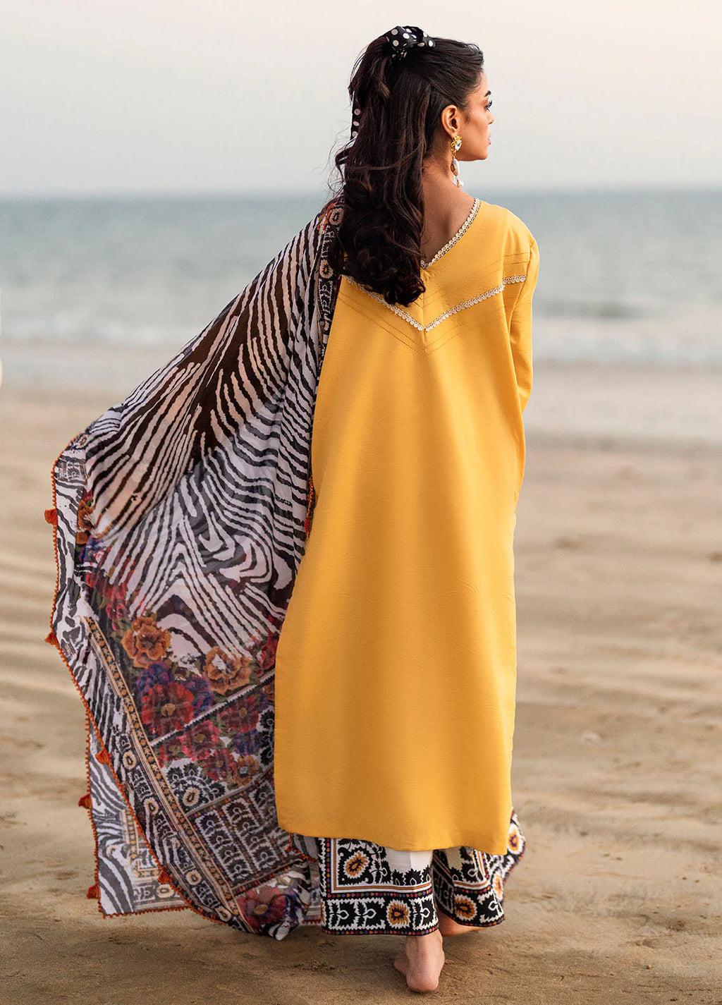 A woman stands on a sandy beach facing away from the camera, wearing a bright yellow kurta with a patterned dupatta and matching patterned pants.