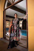 A woman models a traditional embroidered black three-piece outfit with intricate multicolor floral patterns, standing in a doorway with a swimming pool and warm-toned interior visible in the background.