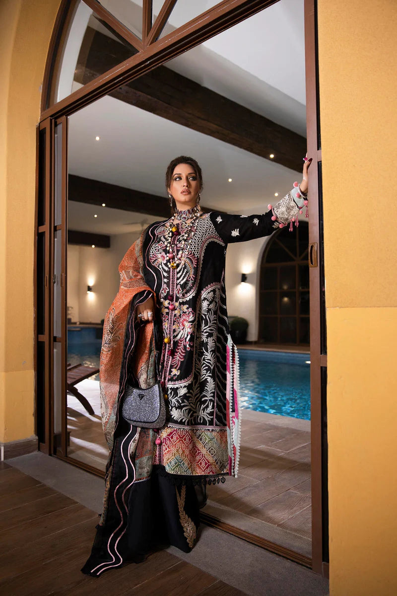 A woman models a traditional embroidered black three-piece outfit with intricate multicolor floral patterns, standing in a doorway with a swimming pool and warm-toned interior visible in the background.