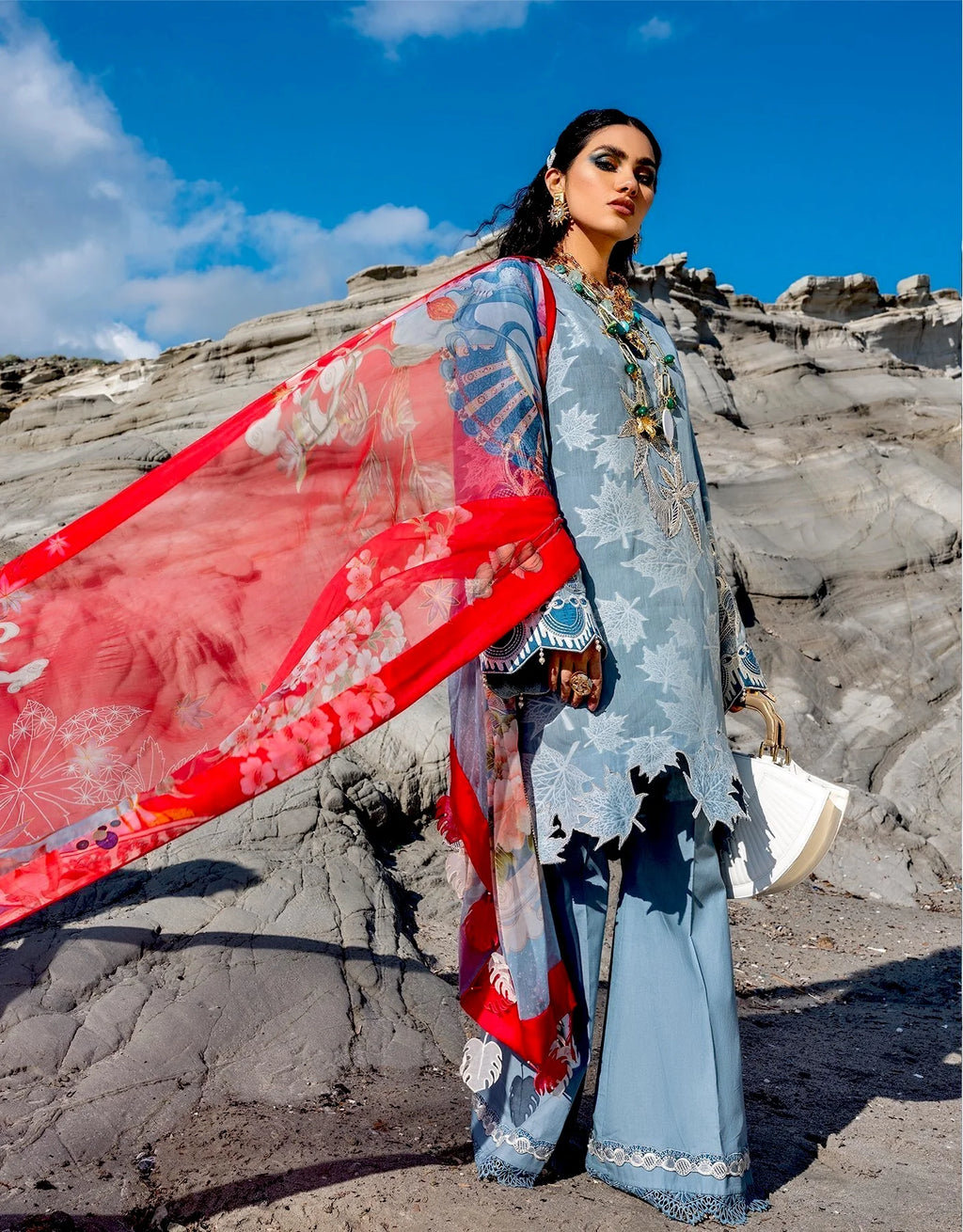 A woman stands outdoors against rocky cliffs under a blue sky, wearing a light blue traditional outfit with a bright red patterned dupatta and holding a white hat.