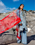 A woman stands outdoors against rocky cliffs under a blue sky, wearing a light blue traditional outfit with a bright red patterned dupatta and holding a white hat.