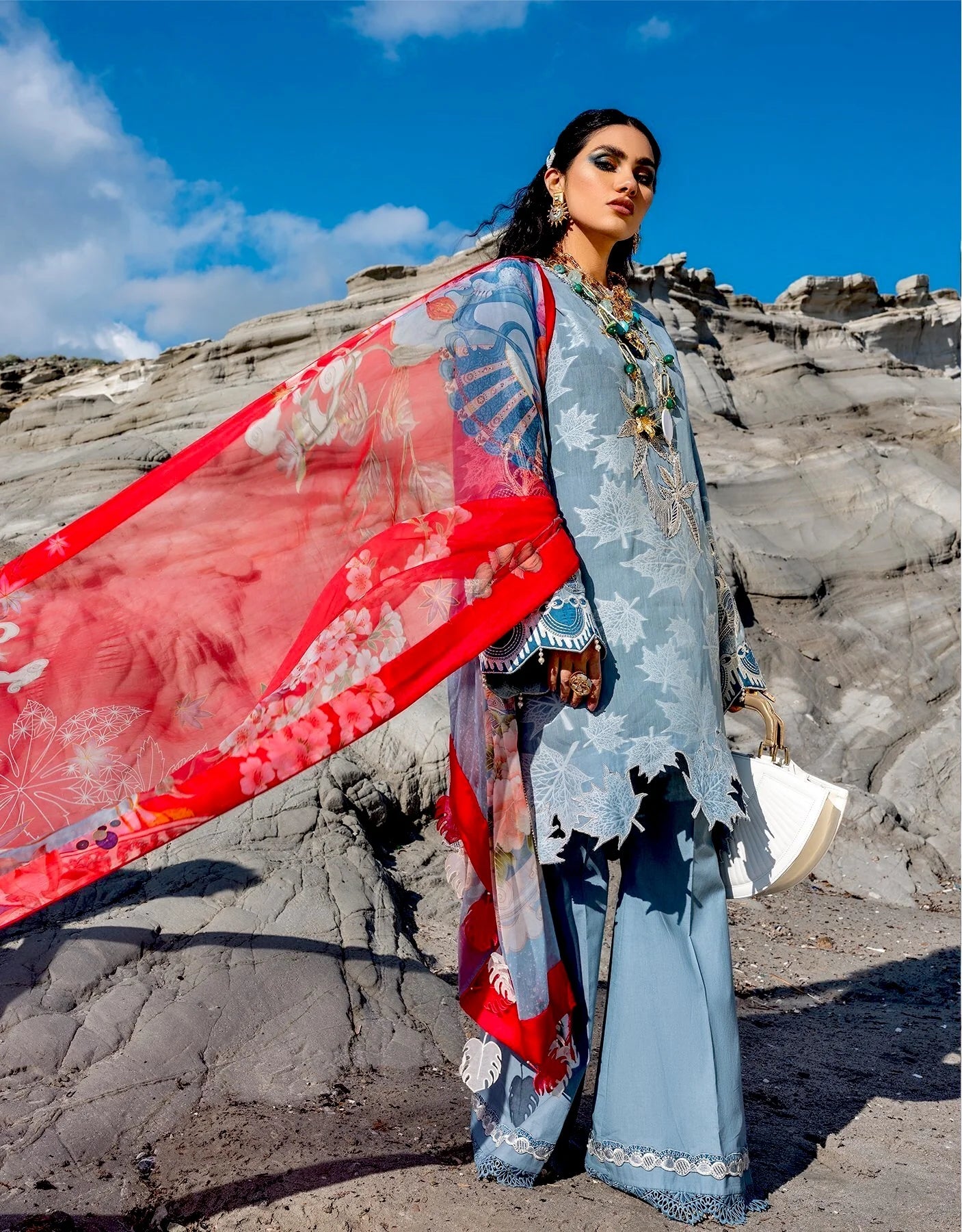 A woman stands outdoors against rocky cliffs under a blue sky, wearing a light blue traditional outfit with a bright red patterned dupatta and holding a white hat.
