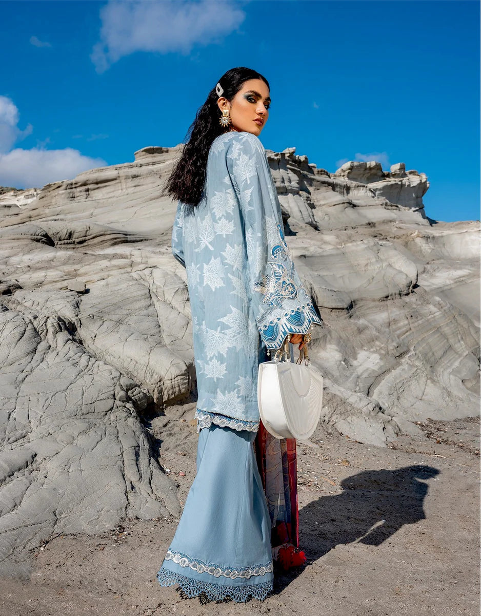 A woman stands outdoors in front of rocky desert formations under a bright blue sky, wearing a light blue traditional outfit with white floral embroidery and holding a white handbag.