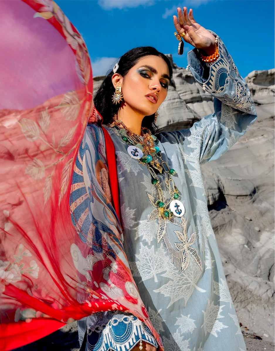 A woman outdoors wearing a light blue embroidered traditional outfit with a red sheer dupatta, layered with ornate gold and turquoise jewelry, against a rocky gray background and blue sky.