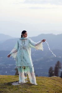 Woman in a light blue traditional outfit standing on a grassy hill with mountains in the background