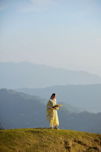 Person in a yellow dress standing on a grassy hill with mountains in the background